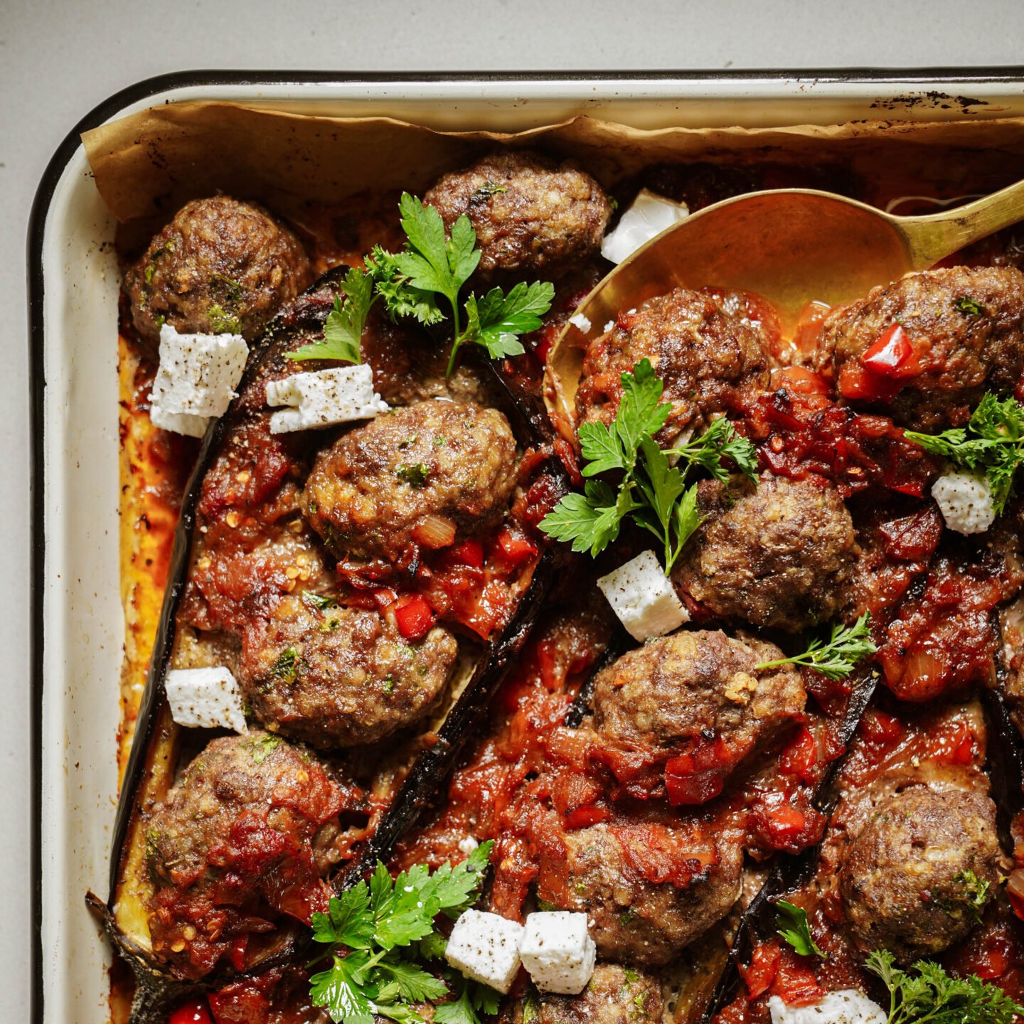 A baking dish filled with roasted eggplant halves topped with meatballs, tomato sauce, fresh parsley, and cubes of white cheese, with a serving spoon on the side.