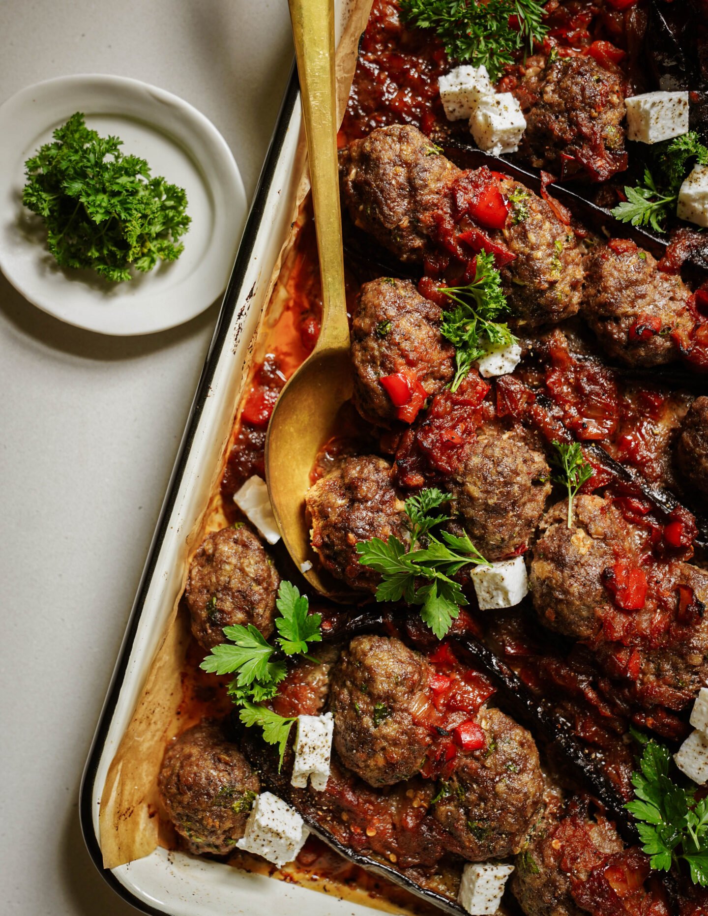 A baking tray of baked meatballs in tomato sauce, garnished with fresh parsley, diced feta cheese, and red peppers. A gold serving spoon rests on the tray, and a small plate with parsley sits beside it.