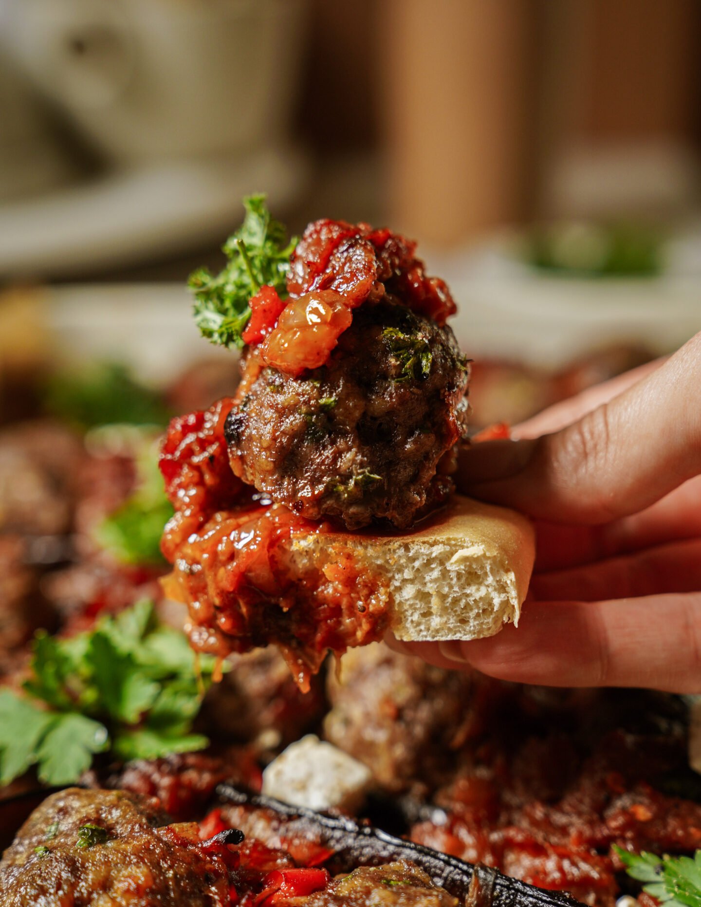 A hand holds a piece of bread topped with a meatball, tomato sauce, and a sprig of parsley. More meatballs and sauce are blurred in the background.