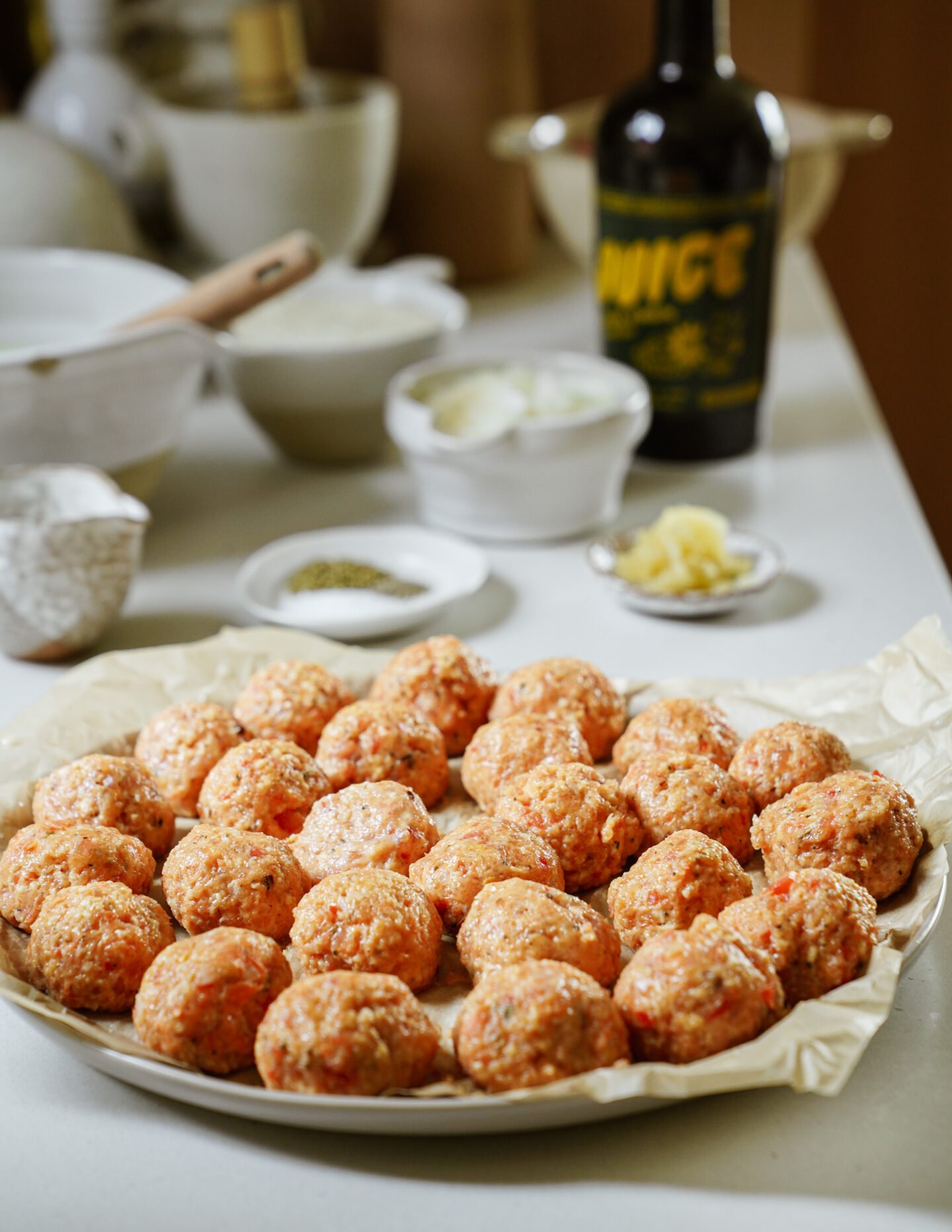 A plate of uncooked meatballs sits on parchment paper. In the background, there are bowls of ingredients, a bottle of olive oil, and a wooden spoon on a kitchen countertop.