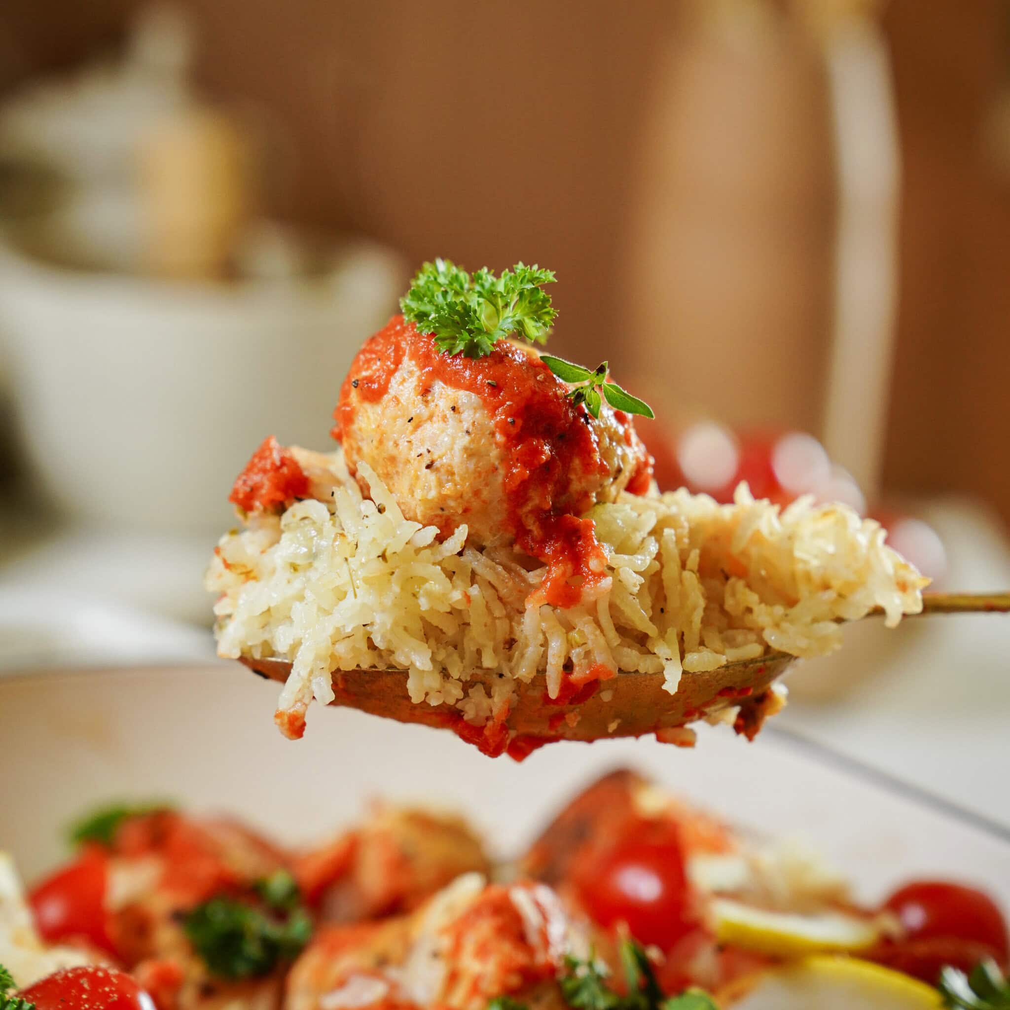 A close-up of a spoonful of rice and a meatball topped with tomato sauce and a parsley garnish, with more rice, meatballs, and vegetables visible in the blurred background.
