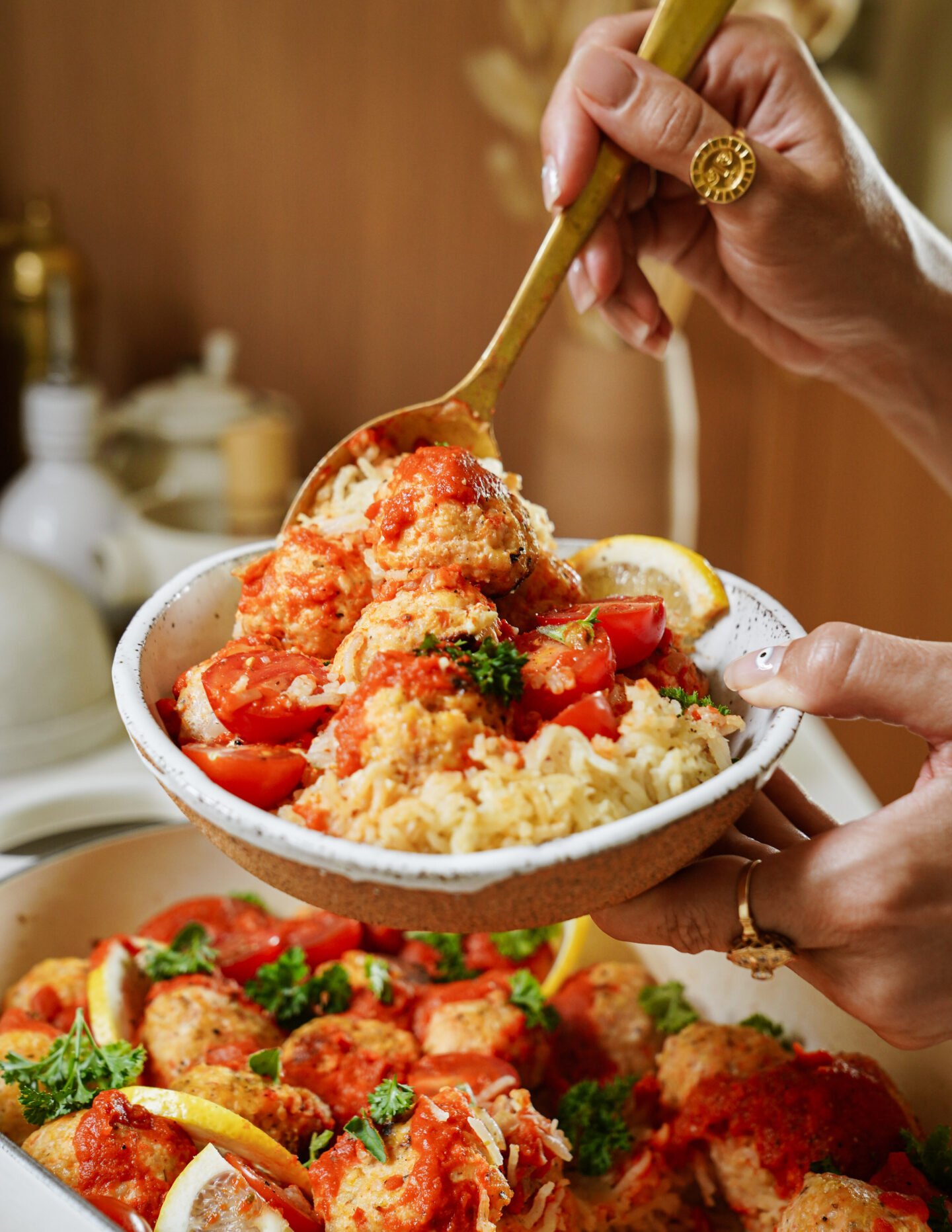A hand holds a bowl of rice topped with tomato sauce, meatballs, cherry tomatoes, and parsley, while another spoonful is being served from a dish below. Lemon slices are visible as garnish.
