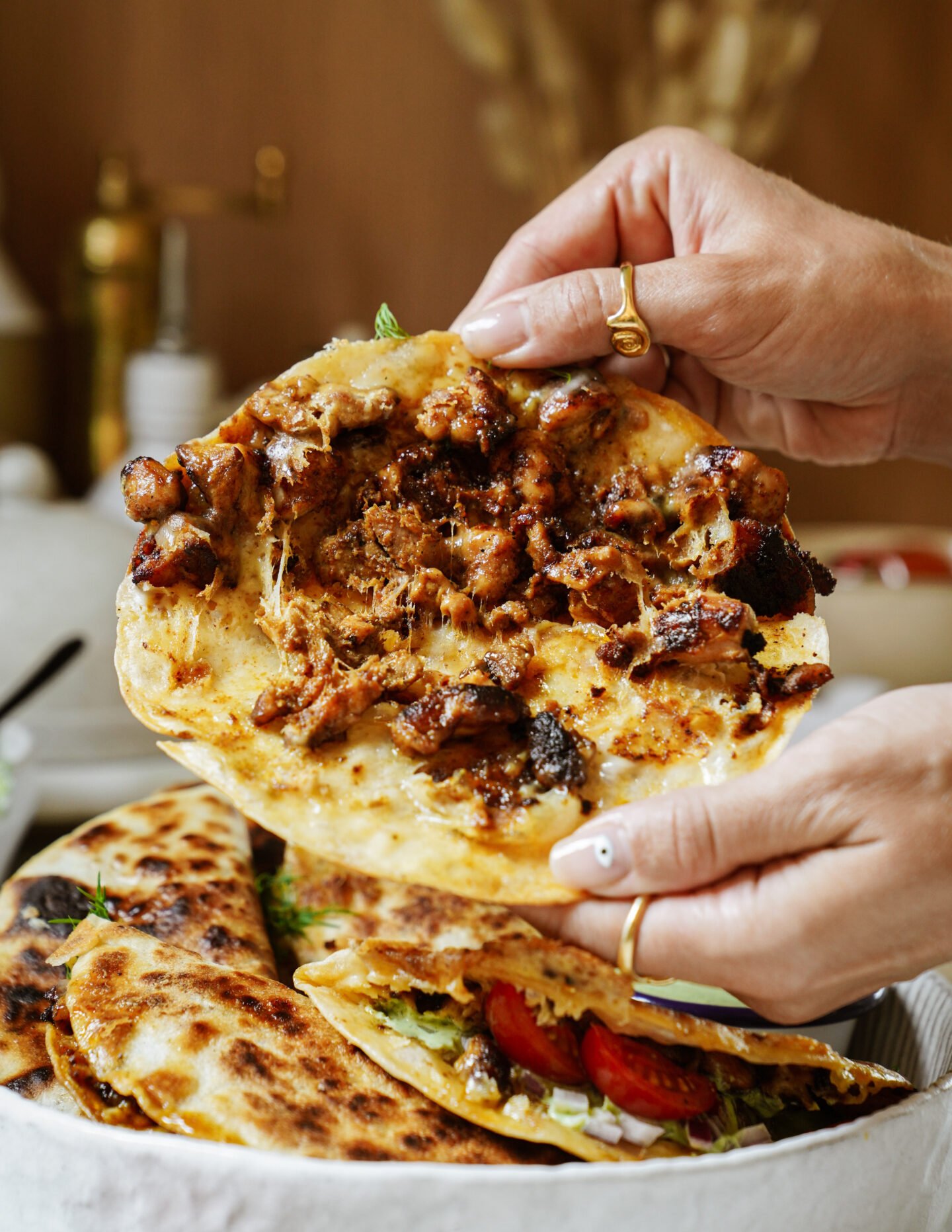 A person holds a toasted flatbread taco filled with seasoned, grilled meat and melted cheese above a dish containing more stuffed flatbreads and fresh vegetables.