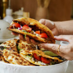 A person holds a folded quesadilla filled with cherry tomatoes, onions, and melted cheese. More quesadillas with similar fillings are on a white plate in the background.
