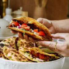 A person holds a folded quesadilla filled with cherry tomatoes, onions, and melted cheese. More quesadillas with similar fillings are on a white plate in the background.