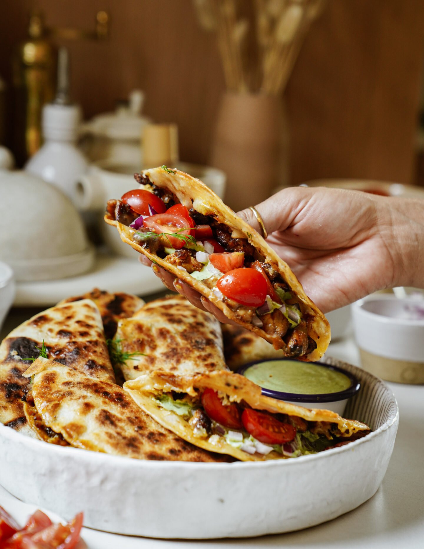 A hand holds a folded tortilla filled with grilled meat, cherry tomatoes, onions, and greens above a plate of similar tacos, accompanied by a small bowl of green dipping sauce.