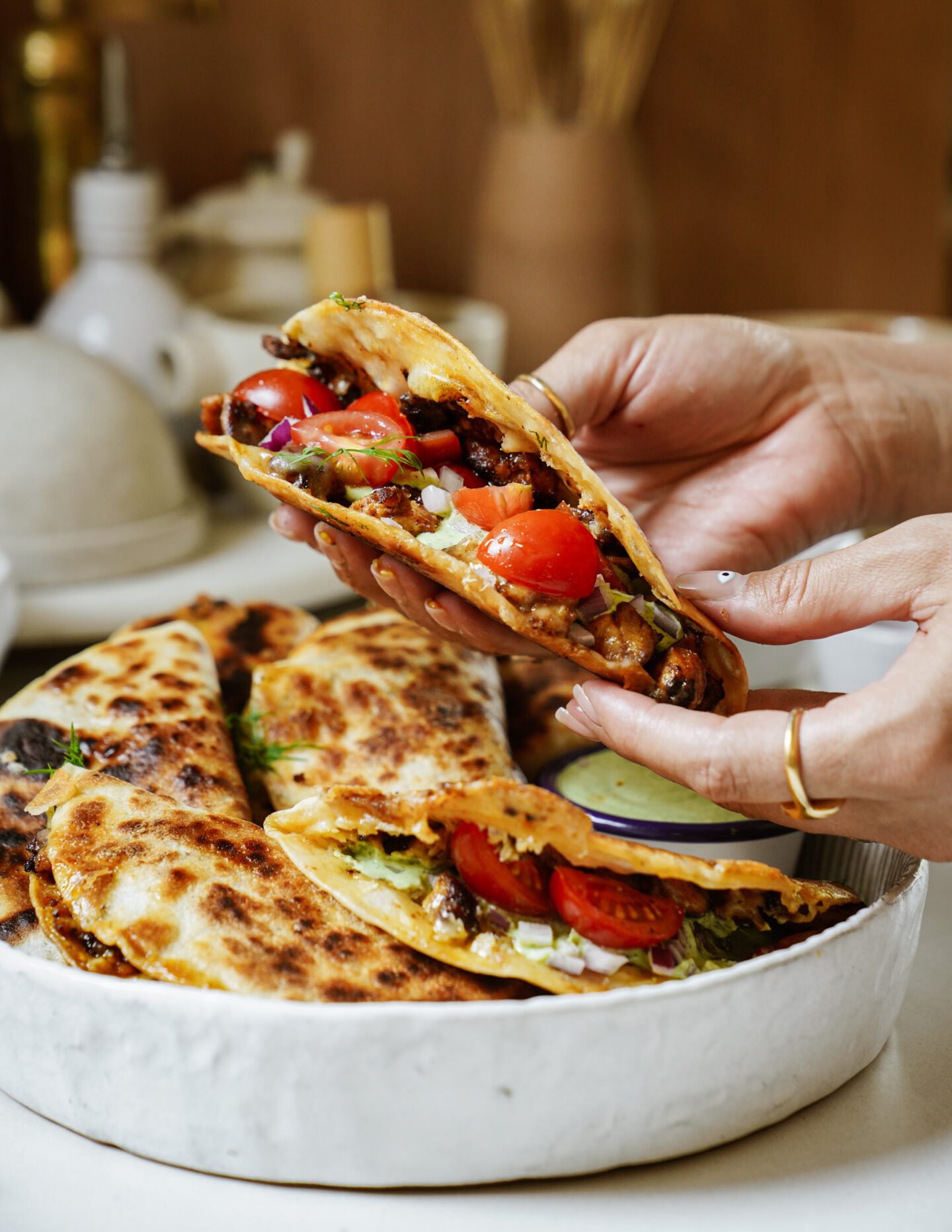 A person holds a folded taco filled with tomatoes, onions, and other vegetables over a white plate with more stuffed and grilled tacos.