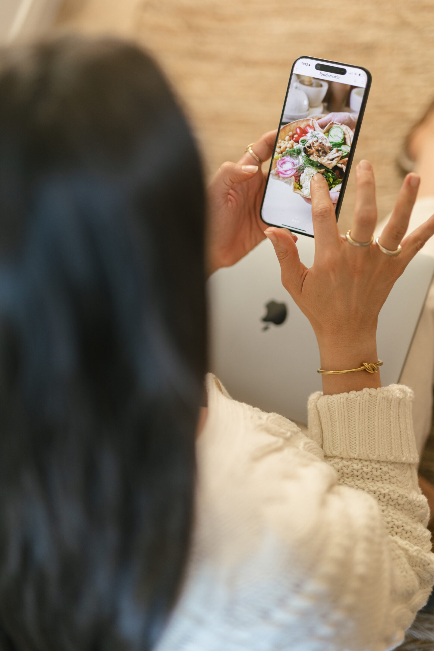 A person with long dark hair holds a smartphone and looks at a photo of food on the screen. They are wearing a cream sweater and gold jewelry, and an Apple laptop is partially visible on their lap.