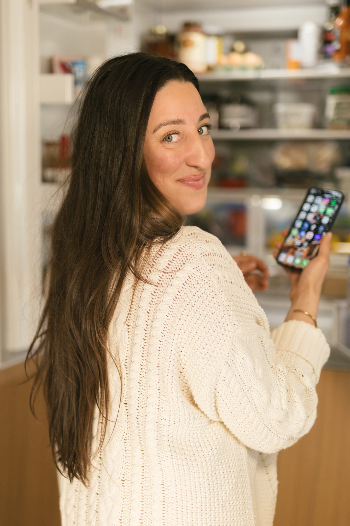 A woman with long brown hair, wearing a cream-colored knitted sweater, stands in front of an open refrigerator, smiling over her shoulder while holding a smartphone with a colorful home screen.