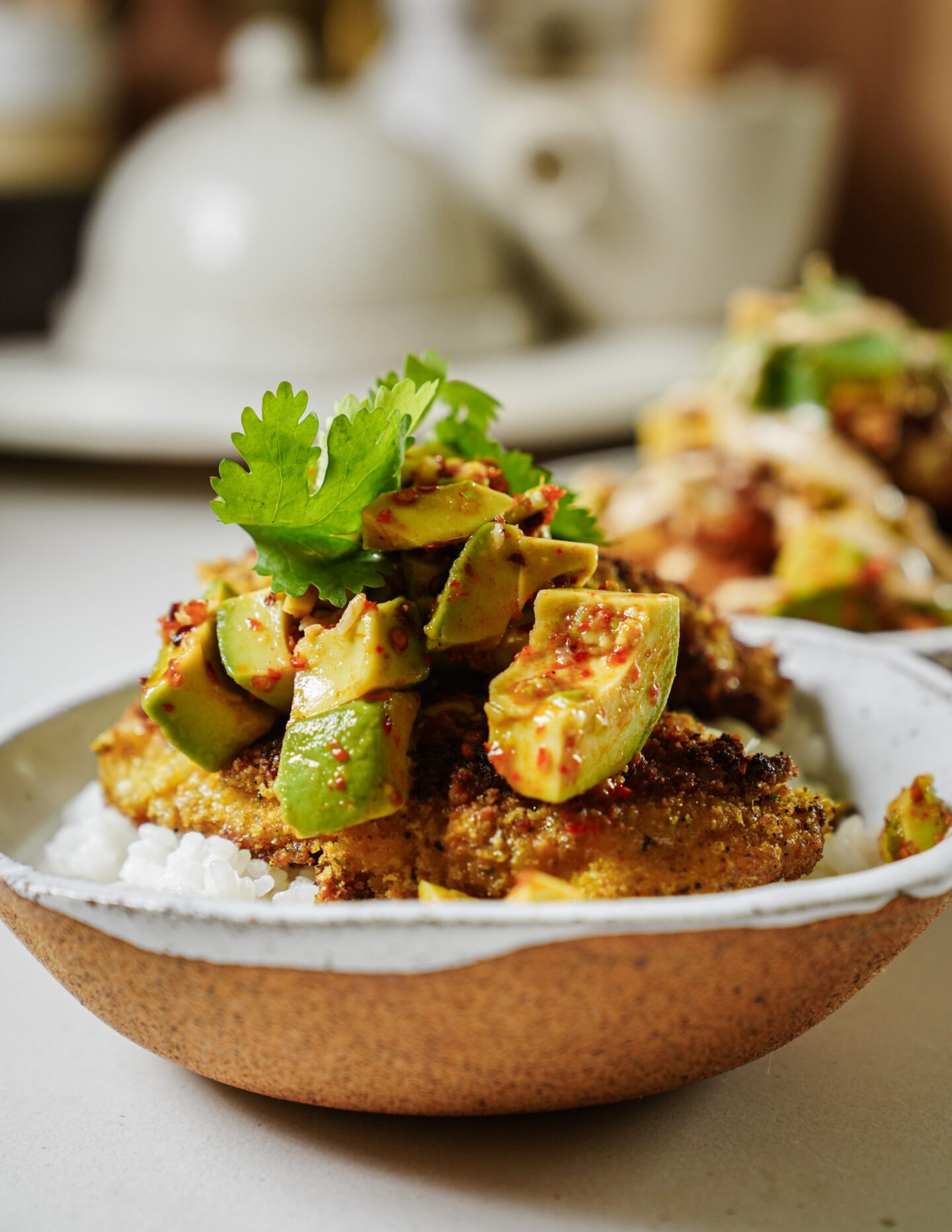 A bowl of white rice topped with a breaded, golden-brown patty, avocado chunks in seasoning, and a sprig of fresh cilantro. The background is softly blurred with tableware visible.