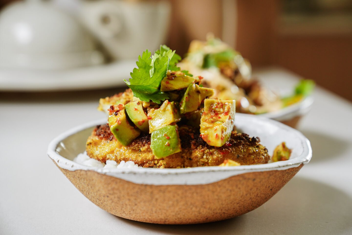 A close-up of a bowl filled with rice, topped with a seasoned patty, diced avocado, herbs, and spices, garnished with a cilantro leaf. Another blurred bowl is visible in the background.
