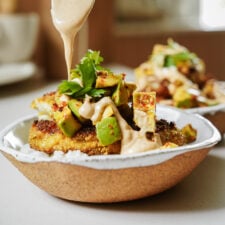 A close-up of a bowl with rice, topped with a crispy patty, avocado salad, and fresh cilantro, as creamy sauce is being drizzled over the dish. Another similar bowl is blurred in the background.