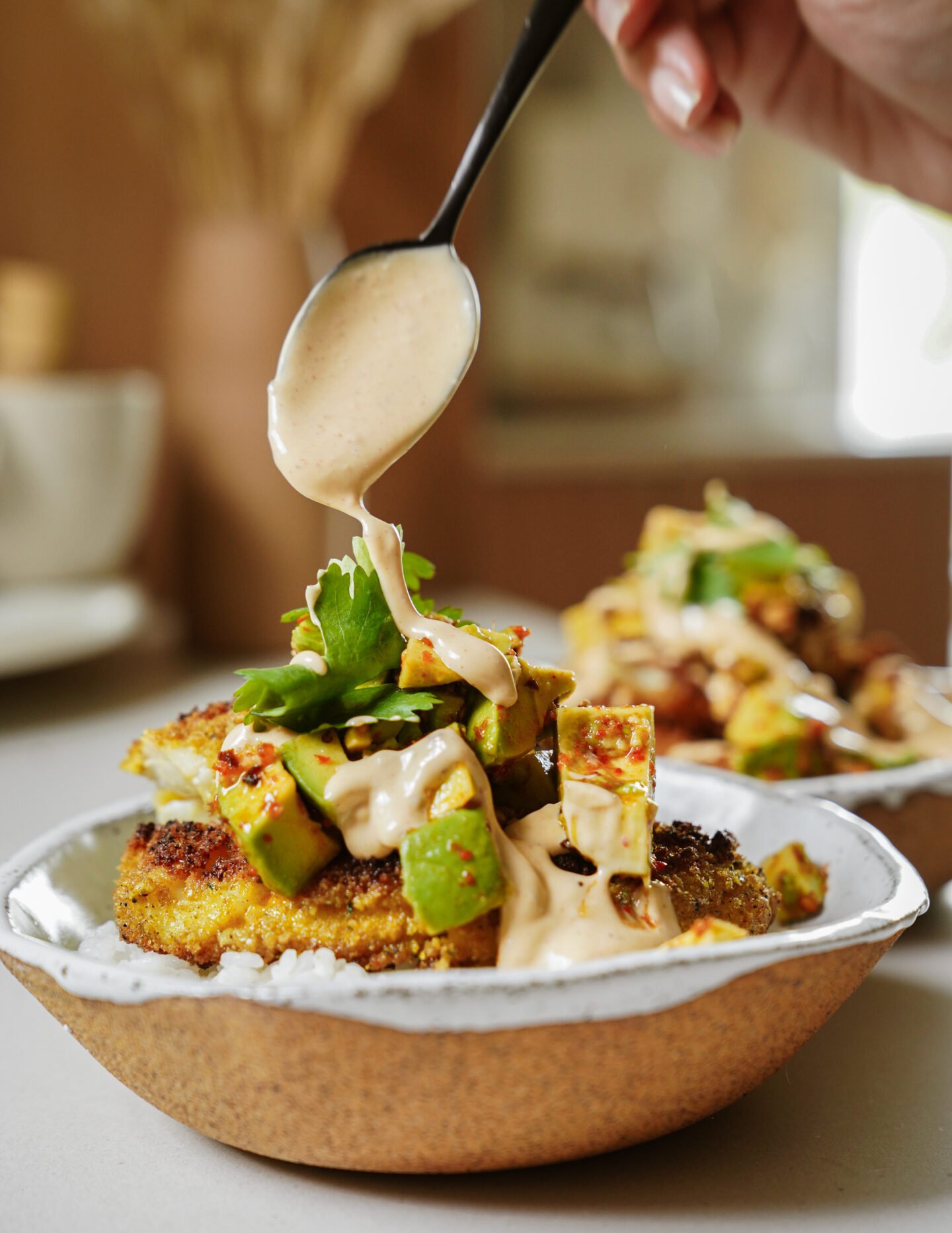 A hand holds a spoon, drizzling creamy sauce over a bowl of rice topped with crispy patties, avocado chunks, and fresh cilantro. Another similar bowl sits blurred in the background.