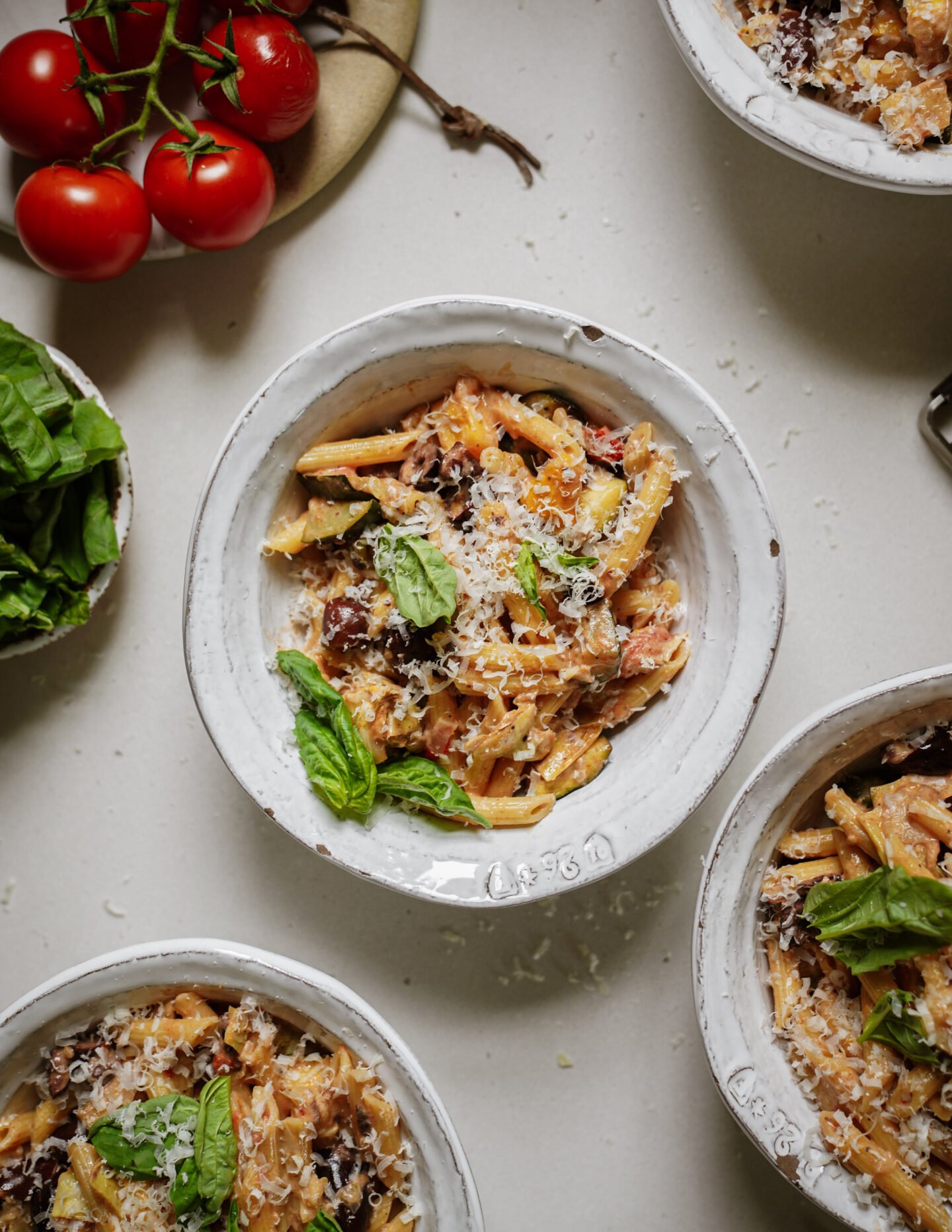 Bowls of pasta primavera topped with grated cheese and fresh basil leaves, surrounded by fresh basil and a cluster of tomatoes on a white surface.