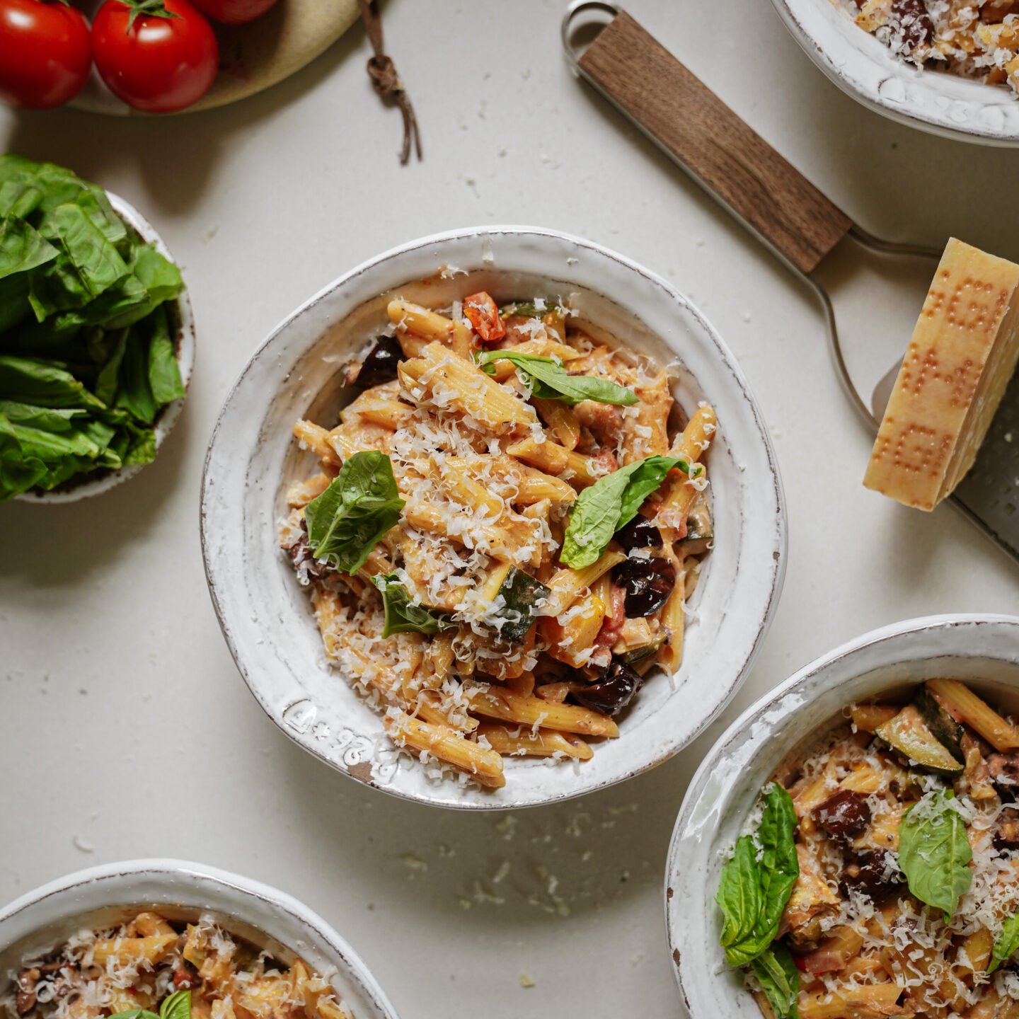 A bowl of pasta topped with grated cheese and fresh basil, surrounded by more bowls of pasta, a block of cheese with a grater, tomatoes on the vine, and a bunch of fresh basil on a white surface.