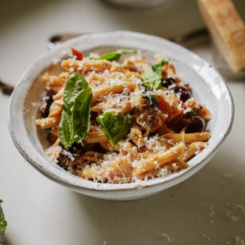 A bowl of pasta topped with grated cheese, fresh basil leaves, and vegetables, placed on a light surface with another bowl of pasta and a wedge of cheese in the background.