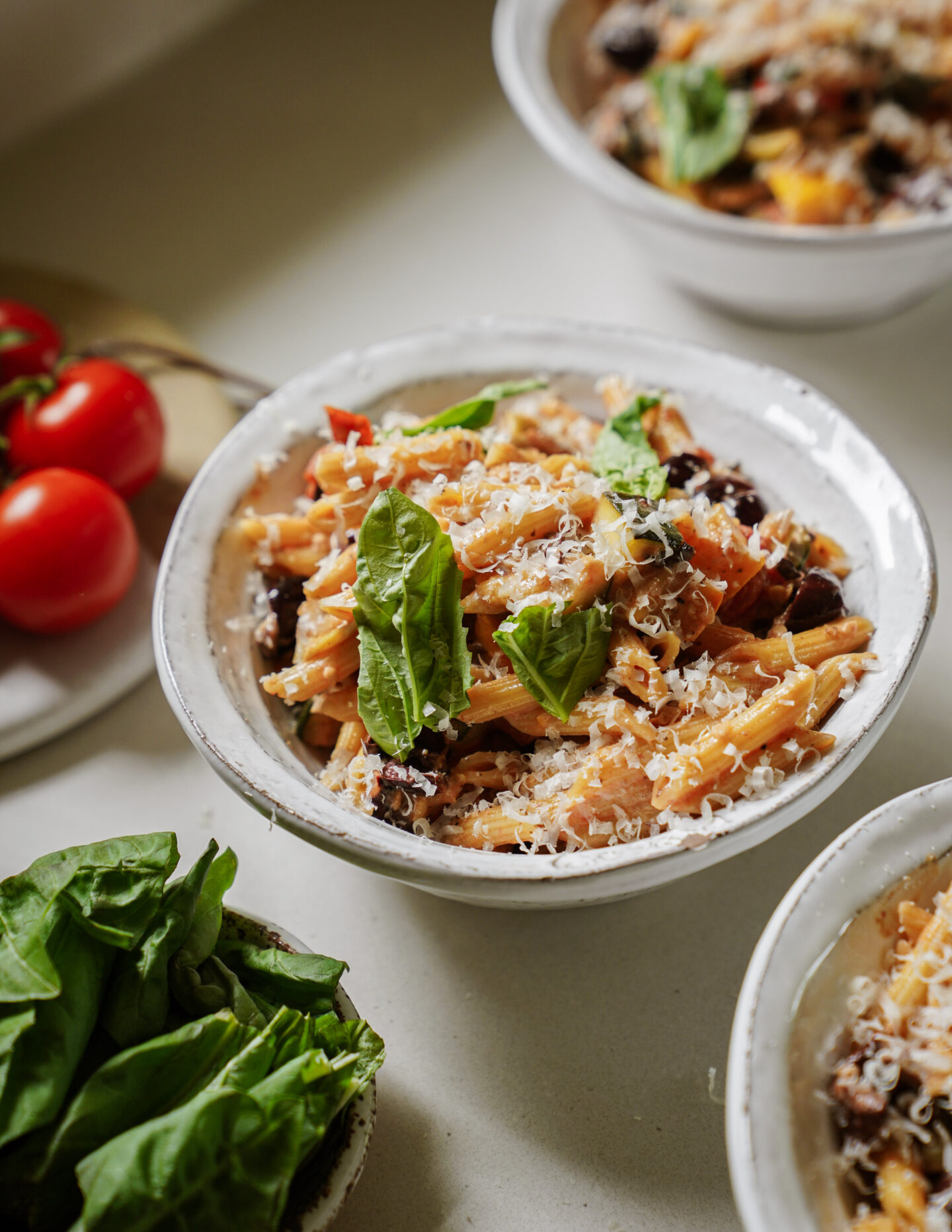 A bowl of pasta topped with grated cheese, fresh basil leaves, and surrounded by more bowls of pasta, cherry tomatoes, and a bowl of fresh basil on a light countertop.