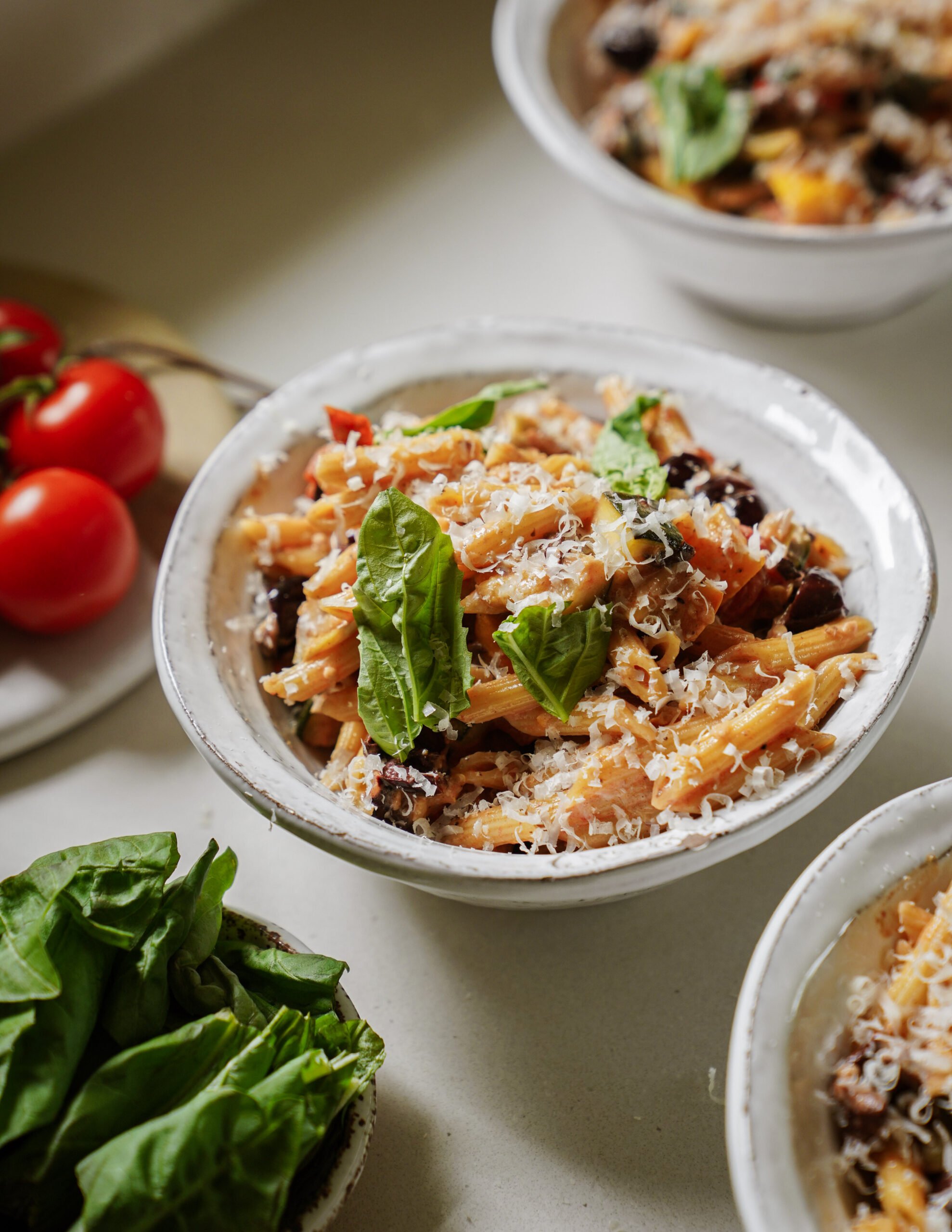 A bowl of pasta topped with grated cheese, fresh basil leaves, and surrounded by more bowls of pasta, cherry tomatoes, and a bowl of fresh basil on a light countertop.