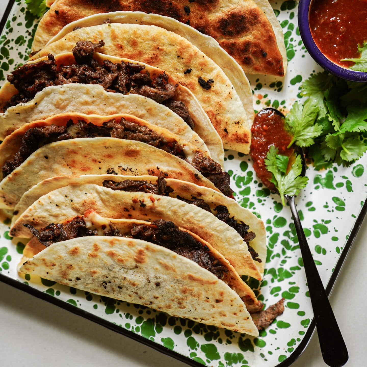Five folded beef tacos on a speckled green and white tray, served with a small bowl of red salsa, a spoon, and fresh cilantro garnish.