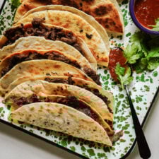 Five folded beef tacos on a speckled green and white tray, served with a small bowl of red salsa, a spoon, and fresh cilantro garnish.