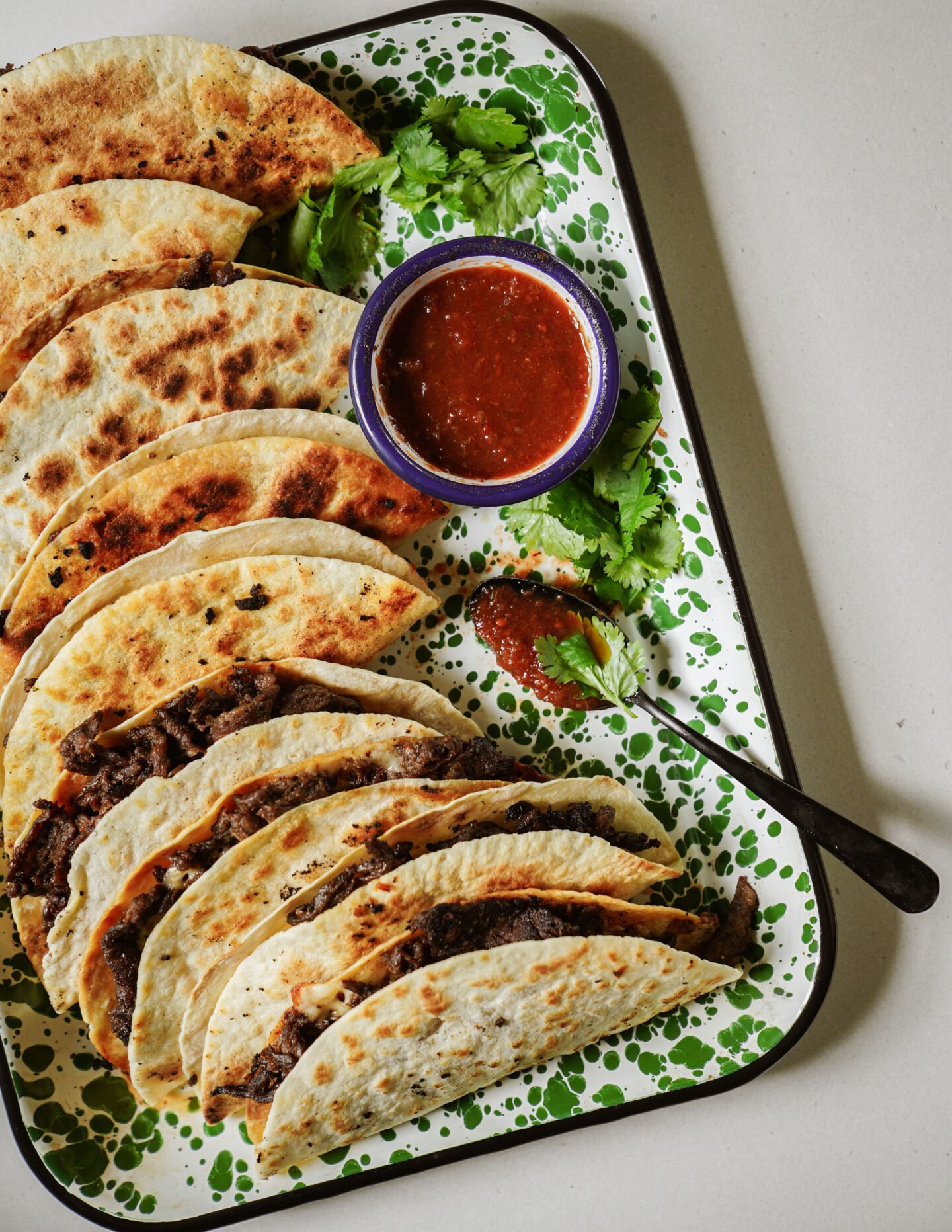 A tray of folded, grilled tacos filled with shredded beef, garnished with fresh cilantro. A small bowl of red salsa and a spoon are on the tray. The tray has a green and white splatter pattern.