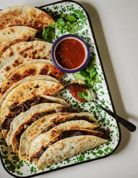 A tray of folded, grilled tacos filled with shredded beef, garnished with fresh cilantro. A small bowl of red salsa and a spoon are on the tray. The tray has a green and white splatter pattern.