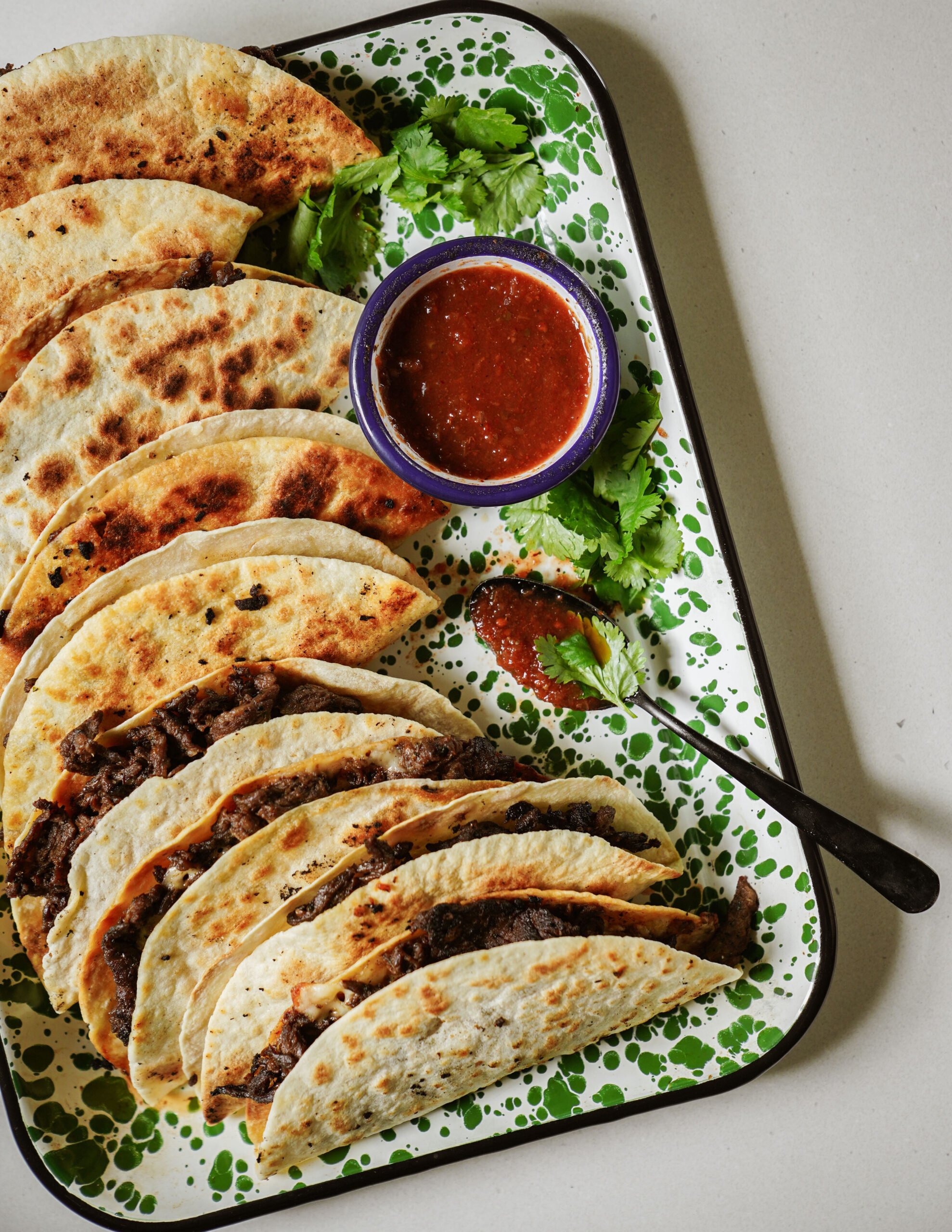 A tray of folded, grilled tacos filled with shredded beef, garnished with fresh cilantro. A small bowl of red salsa and a spoon are on the tray. The tray has a green and white splatter pattern.