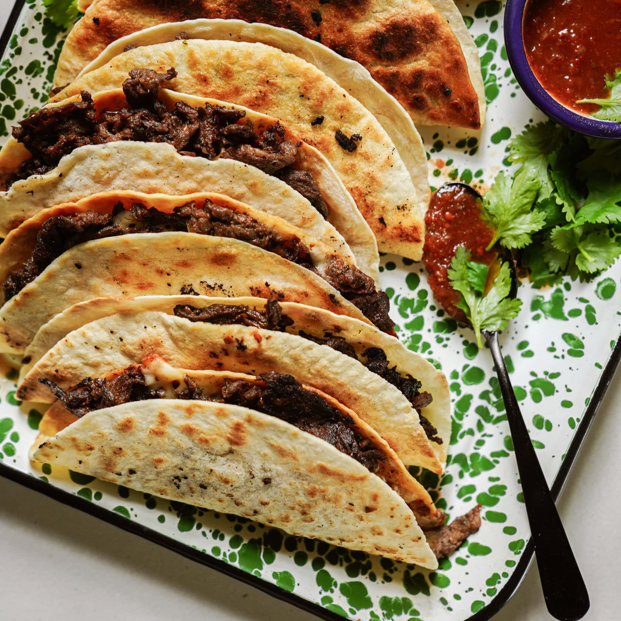 Five folded beef tacos on a speckled green and white tray, served with a small bowl of red salsa, a spoon, and fresh cilantro garnish.