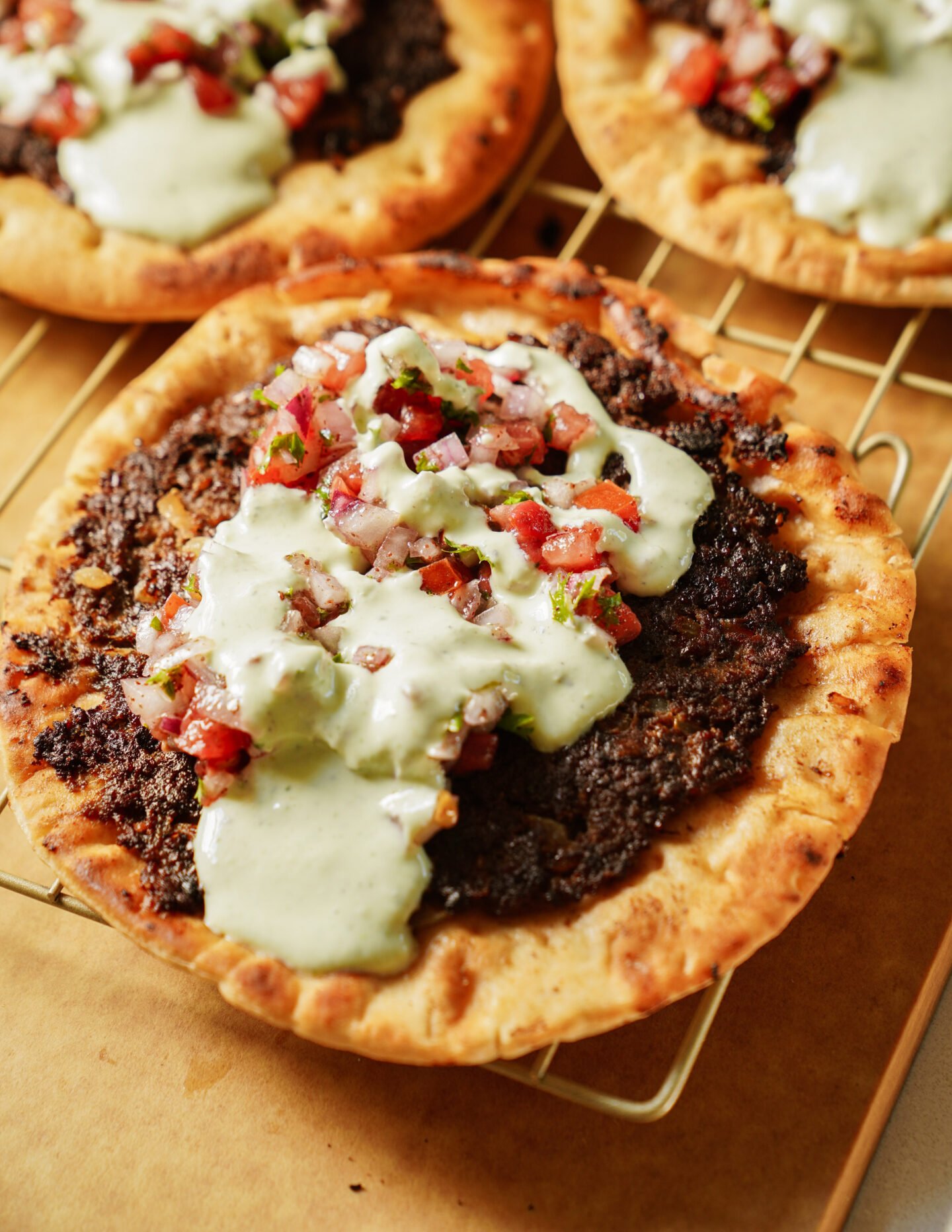 A close-up of a crispy flatbread topped with a dark savory spread, drizzled with creamy white sauce, and garnished with fresh tomato salsa. The flatbread sits on a cooling rack with more flatbreads in the background.