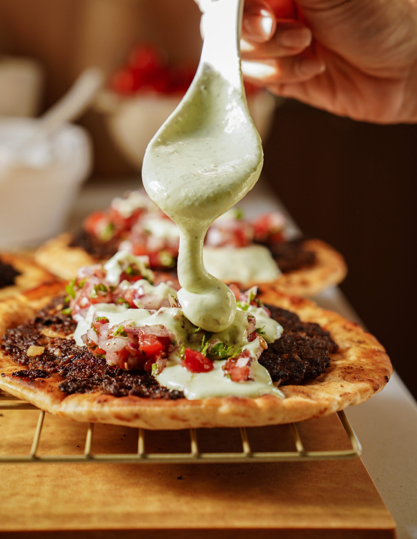 A close-up of a hand pouring creamy green sauce over flatbreads topped with minced meat, tomatoes, and herbs, resting on a wire rack.