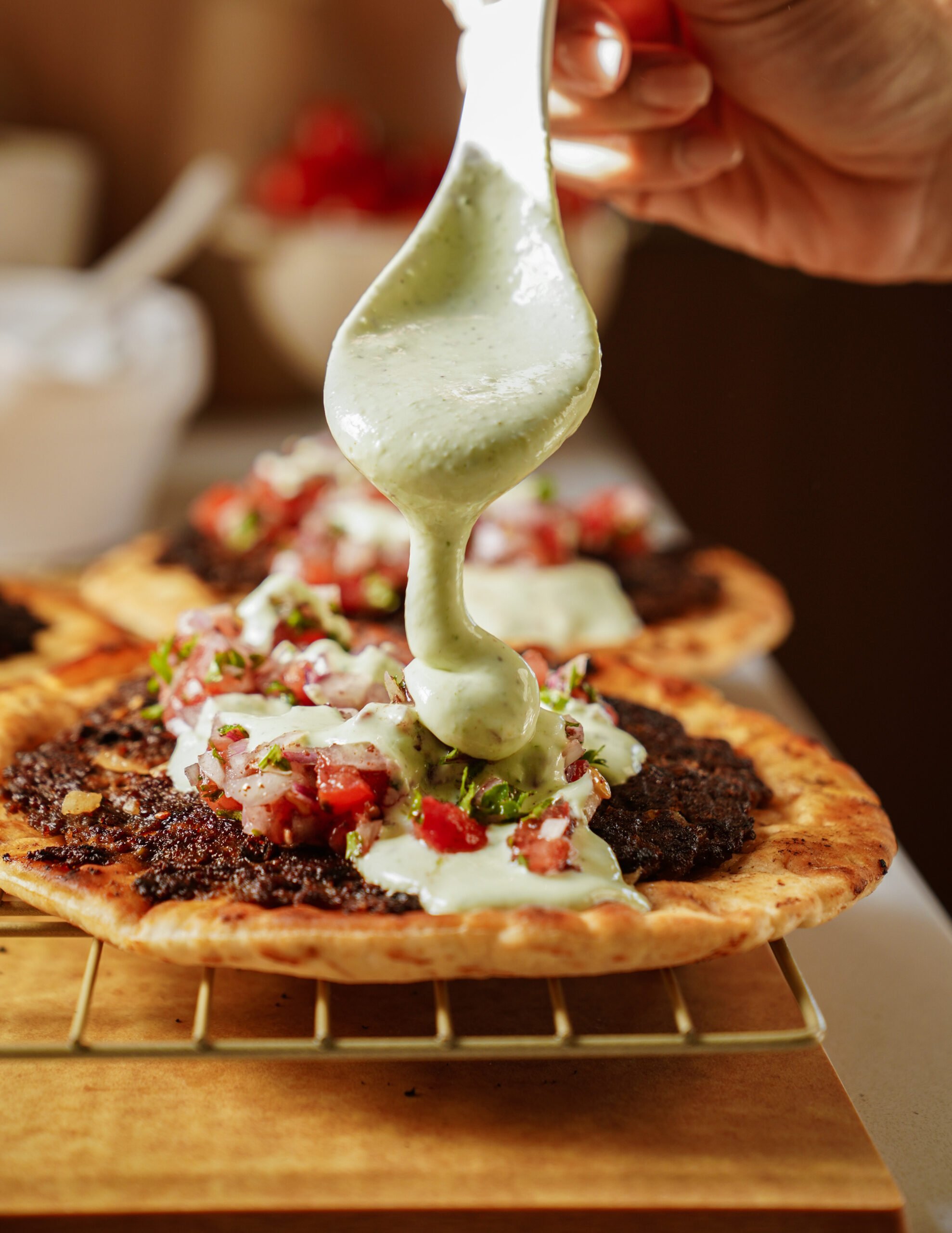 A close-up of a hand pouring creamy green sauce over flatbreads topped with minced meat, tomatoes, and herbs, resting on a wire rack.