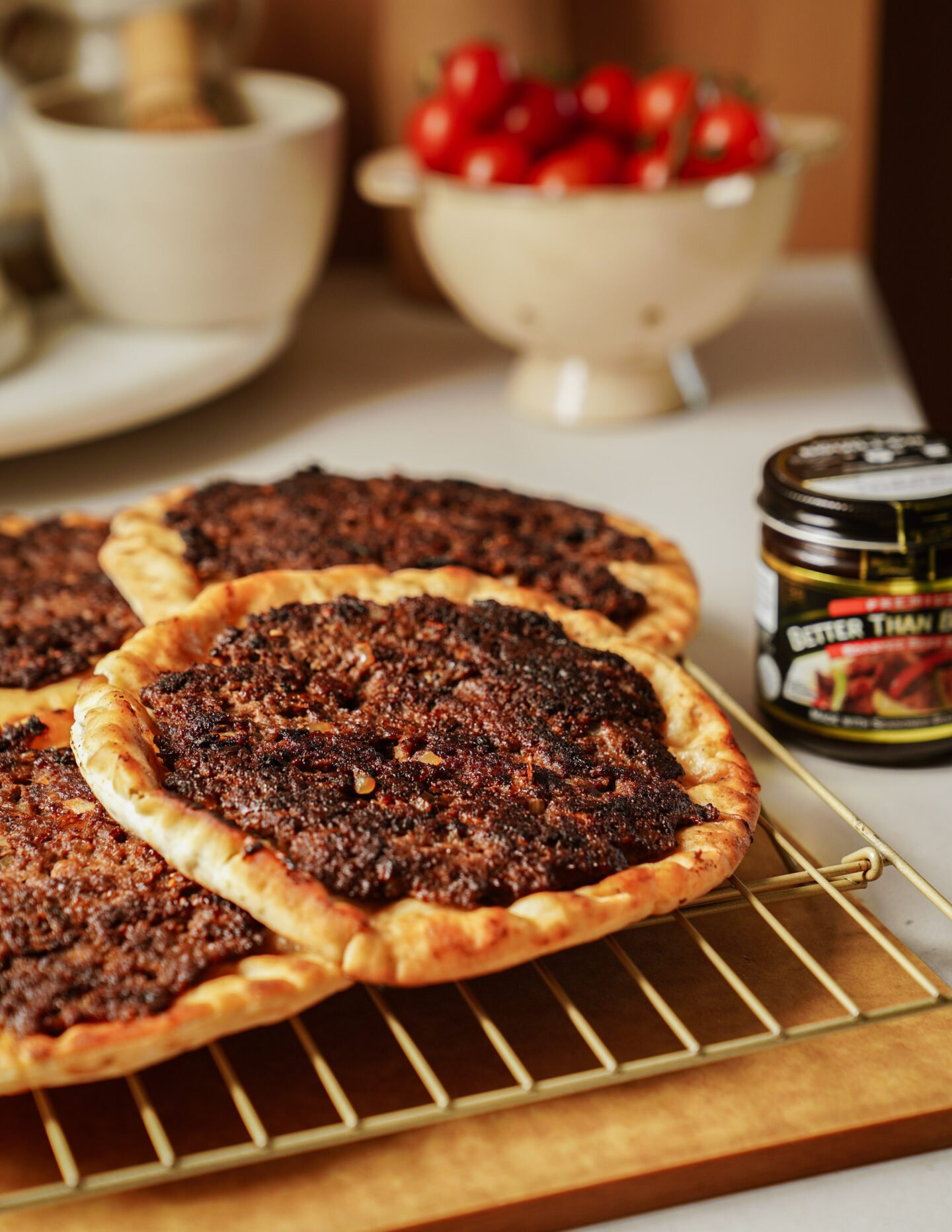 Three baked flatbreads with a dark, spiced topping are cooling on a wire rack. In the background, there is a bowl of cherry tomatoes and a jar labeled Better Than Bouillon.