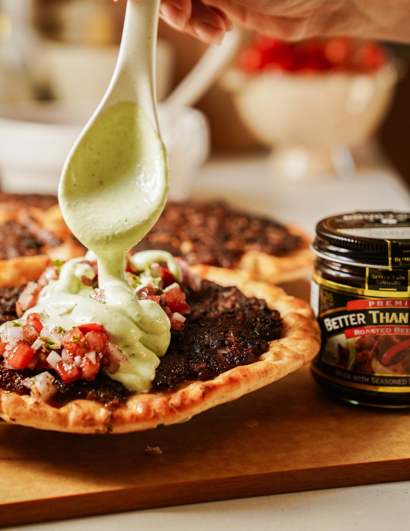 A hand pours a creamy green sauce from a spoon onto a savory pie topped with diced tomatoes and herbs. Another similar pie and a jar of Better Than Bouillon Roasted Beef Base are nearby on the wooden surface.