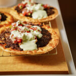 A close-up of two flatbreads topped with spiced ground meat, fresh tomato salsa, and a generous drizzle of creamy white sauce, served on a wire rack over a wooden board.