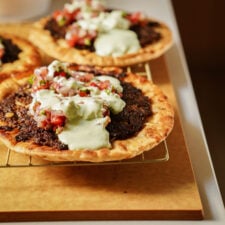 A close-up of two flatbreads topped with spiced ground meat, fresh tomato salsa, and a generous drizzle of creamy white sauce, served on a wire rack over a wooden board.