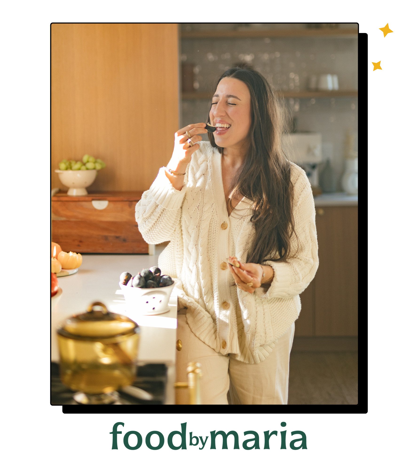 A woman with long dark hair, wearing a cream sweater, stands in a sunlit kitchen enjoying a snack. She is smiling with her eyes closed, holding food in one hand. The text food by maria appears at the bottom.