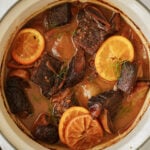 A pot of braised beef short ribs in a rich brown sauce, garnished with slices of orange and fresh herbs. The meat appears tender and well-cooked, surrounded by aromatic broth.