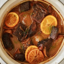 A pot of braised beef short ribs in a rich brown sauce, garnished with slices of orange and fresh herbs. The meat appears tender and well-cooked, surrounded by aromatic broth.