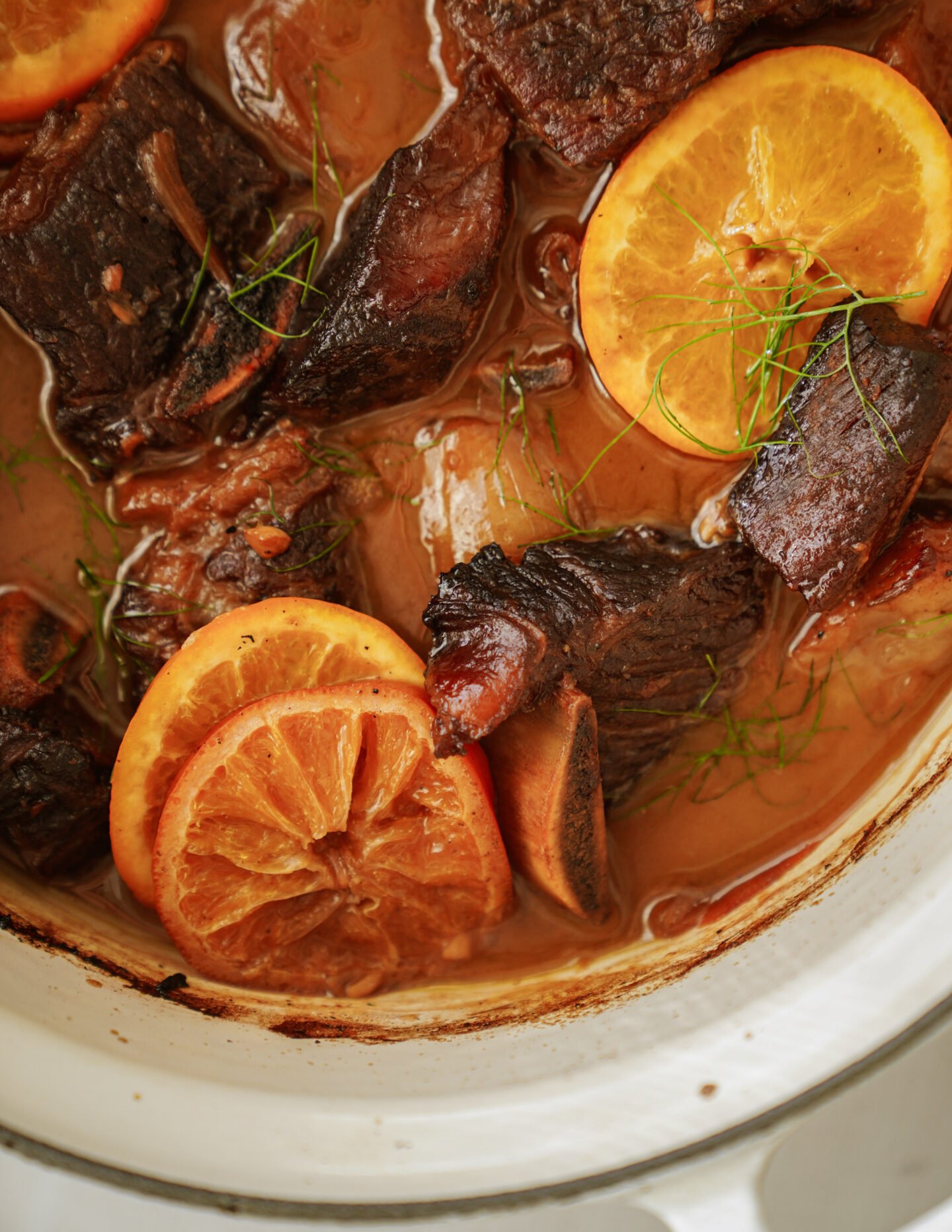 Chunks of braised meat and several orange slices in a rich brown sauce, garnished with fresh green herbs, inside a white pot.