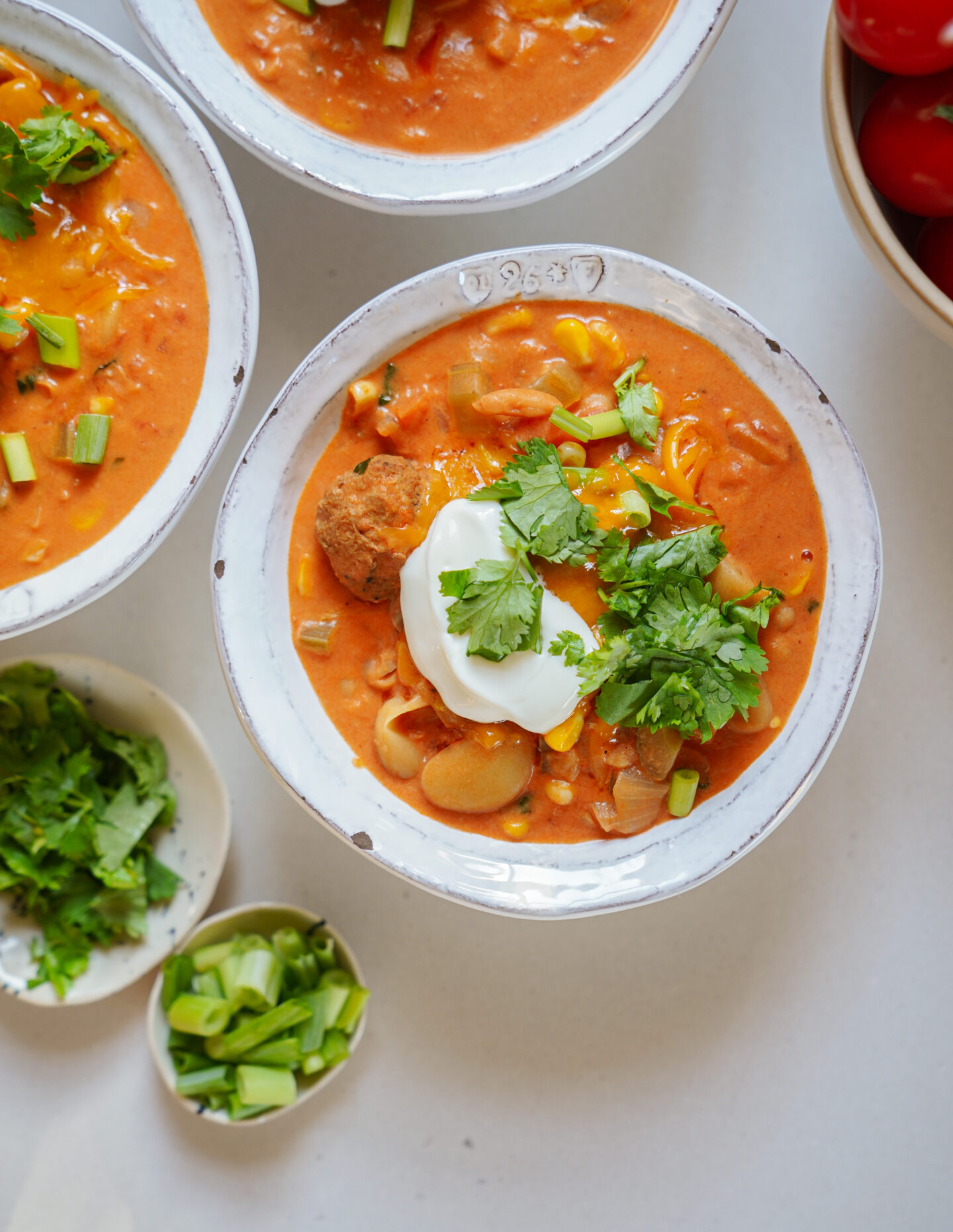 A bowl of chili topped with sour cream, shredded cheese, chopped cilantro, and green onions, surrounded by more bowls of chili and small dishes of fresh cilantro and green onions on a white surface.