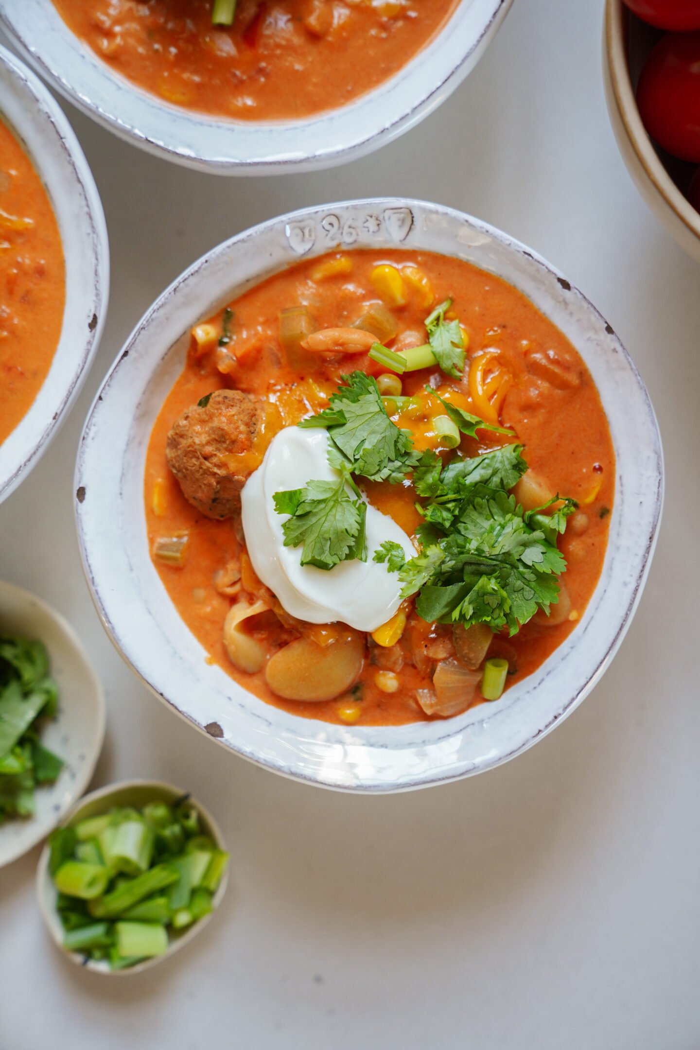 A bowl of creamy tomato-based soup topped with sour cream, fresh cilantro, green onions, and a meatball, surrounded by bowls of chopped green onions and herbs.