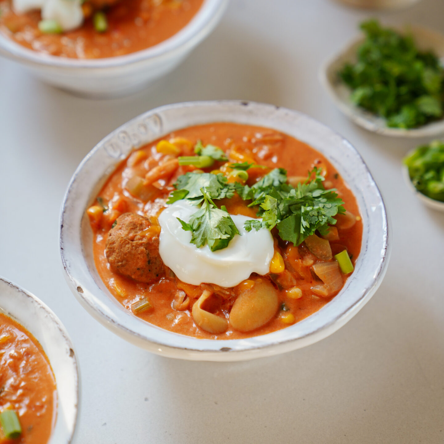 A bowl of creamy tomato soup topped with a dollop of sour cream and fresh cilantro, with visible vegetables and meatballs. Small bowls of chopped herbs are in the background.