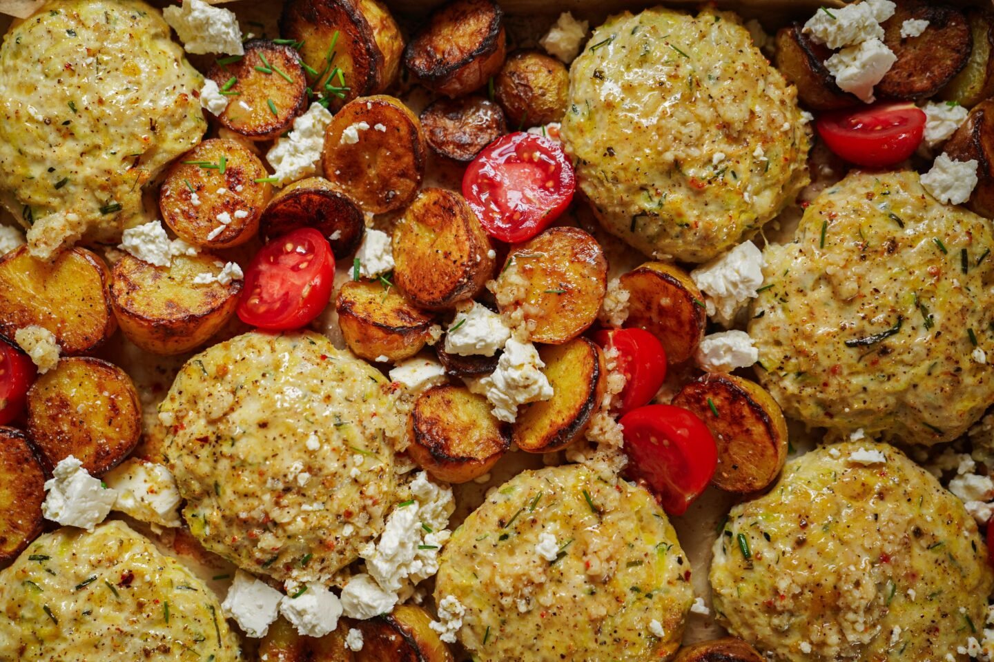 Golden chicken patties, roasted sliced potatoes, crumbled feta cheese, and halved cherry tomatoes arranged together on a tray.