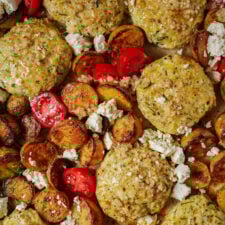 Golden-brown roasted potatoes, zucchini patties, cherry tomato halves, and crumbled feta cheese are arranged closely together on a baking sheet. The dish looks colorful and freshly prepared.