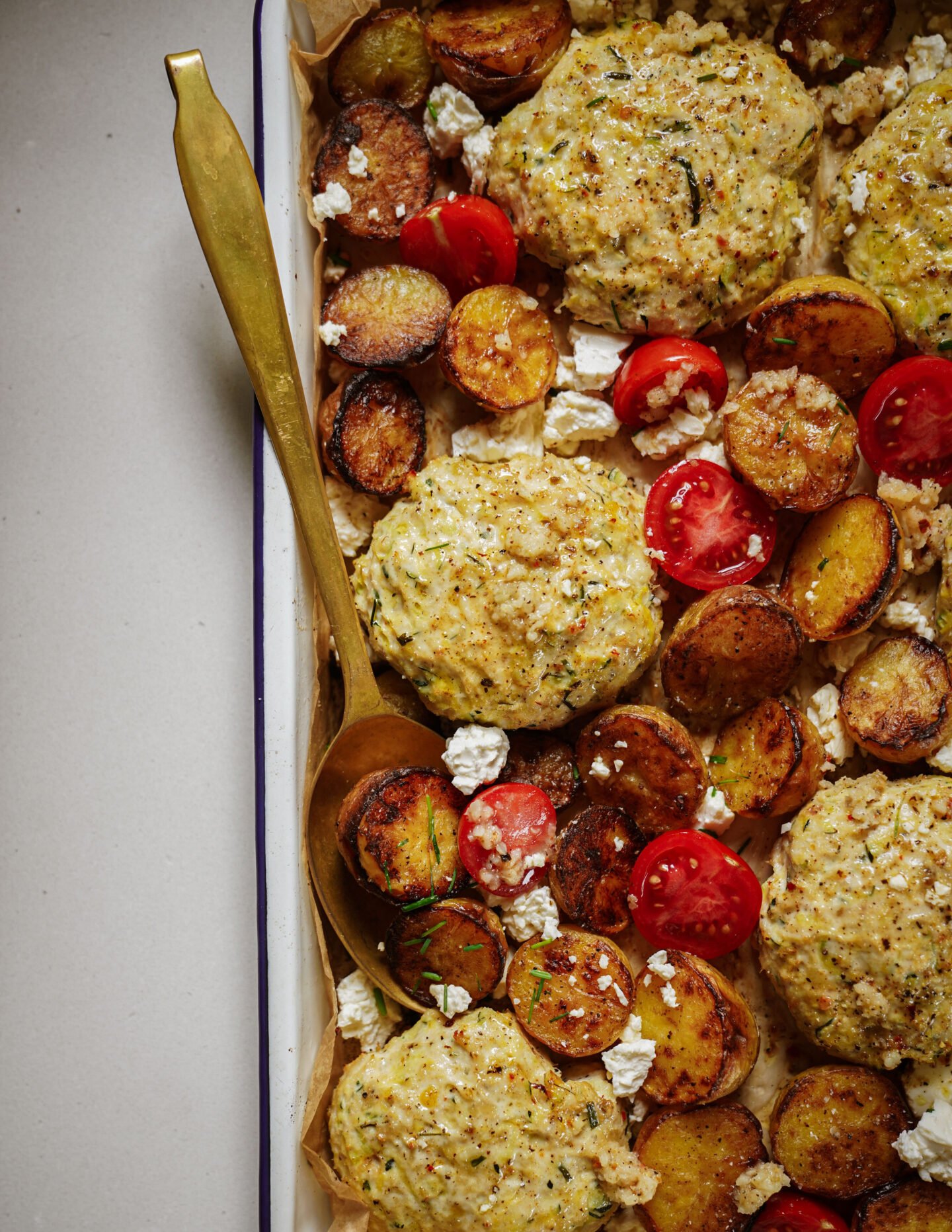 Baked chicken meatballs, roasted sliced potatoes, halved cherry tomatoes, and crumbled feta cheese on a parchment-lined tray with a gold serving spoon.