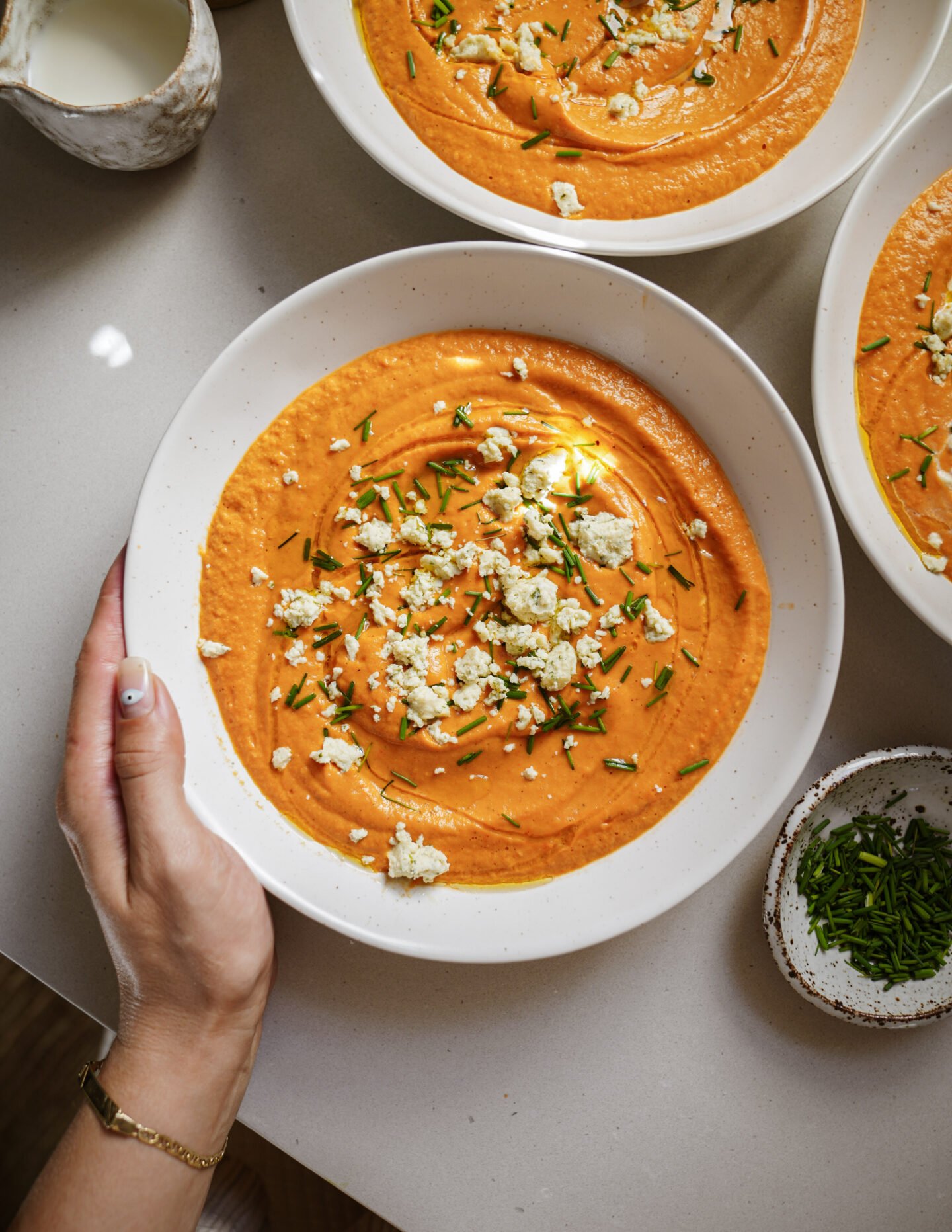 A hand holds a bowl of creamy orange soup topped with crumbled cheese and chopped chives. More bowls of soup, a small bowl of chives, and a jug of milk are on the light-colored tabletop.