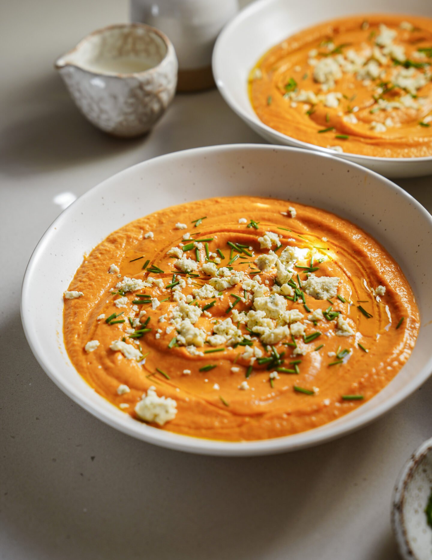 A white bowl filled with creamy orange soup, topped with crumbled cheese and chopped herbs, sits on a light surface. Another similar bowl and a small ceramic pitcher are in the background.