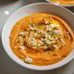 A bowl of creamy orange dip, possibly roasted red pepper hummus, garnished with crumbled cheese, chopped chives, and a drizzle of olive oil on top.