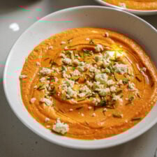 A bowl of creamy orange dip, possibly roasted red pepper hummus, garnished with crumbled cheese, chopped chives, and a drizzle of olive oil on top.
