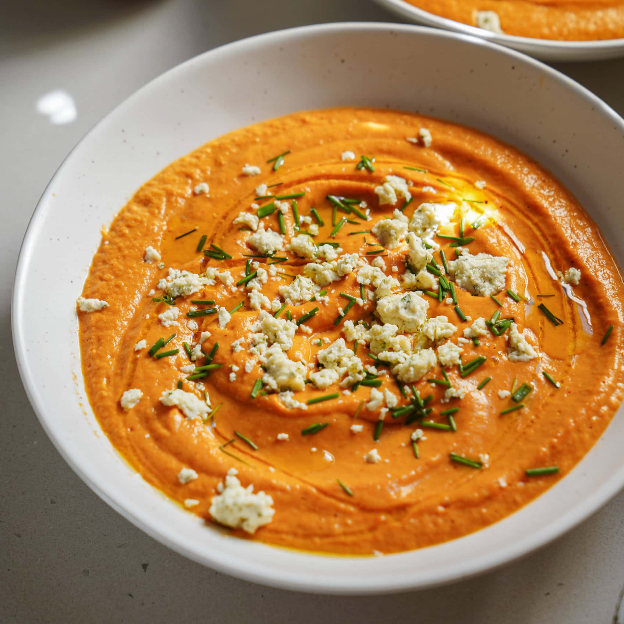 A bowl of creamy orange dip, possibly roasted red pepper hummus, garnished with crumbled cheese, chopped chives, and a drizzle of olive oil on top.